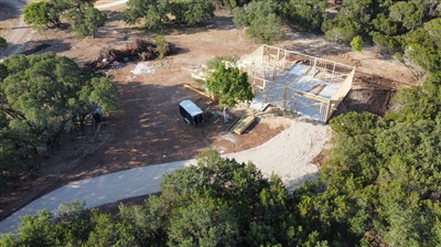 Aerial view of a construction site in a wooded area with a partially built structure, a truck, and a trailer.