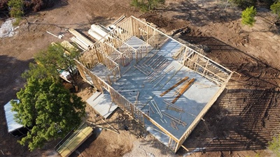 Aerial view of a partially constructed building with visible wooden framework and concrete foundation in a cleared area surrounded by trees.