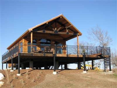 A raised wooden cabin with a large deck and metal railings, situated on a hill with a clear blue sky in the background.