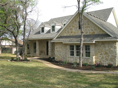 A stone-clad house with a sloped roof and dormer windows, surrounded by a grassy yard and trees.