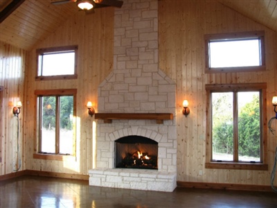 A cozy living room with wooden walls, large windows, and a stone fireplace, illuminated by wall-mounted sconces.