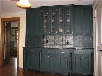 A kitchen with dark green cabinetry and a tiled backsplash, featuring multiple drawers and glass-front upper cabinets.