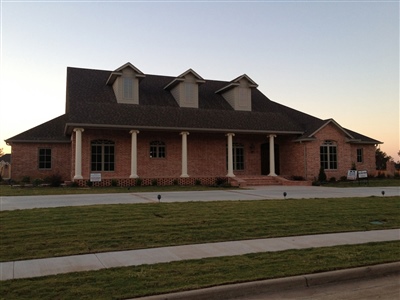 A large brick house with a sloped roof, multiple dormer windows, and a front porch supported by columns, set against a sunset sky.