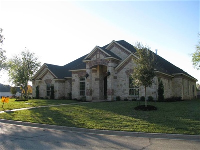 A stone house with a dark sloped roof, manicured lawn, and trees, located at a street corner under a clear sky.