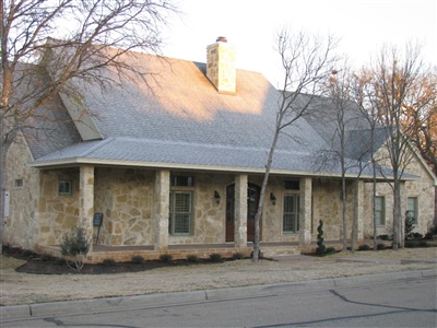 A stone house with a metal roof, front porch supported by columns, and bare trees in the front yard.
